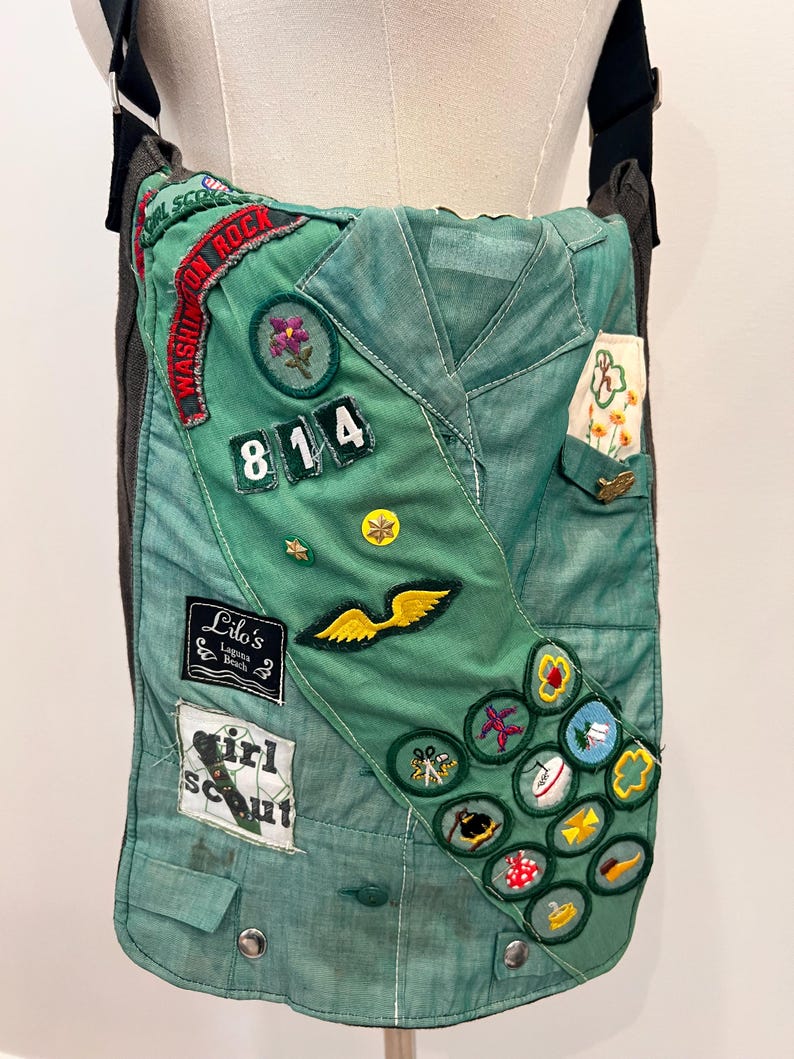 Puede incluir: Un bolso de hombro verde hecho de un uniforme de Girl Scout. El bolso presenta varios parches bordados, incluyendo insignias, un n&uacute;mero de unidad y una etiqueta con el nombre. Tiene una correa negra y un respaldo gris.
