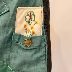 Puede incluir: Primer plano de una tela verde azulado con un bolsillo que presenta un alfiler dorado y un parche bordado con una figura y flores. El parche es blanco con un borde verde. Se ven costuras blancas.
