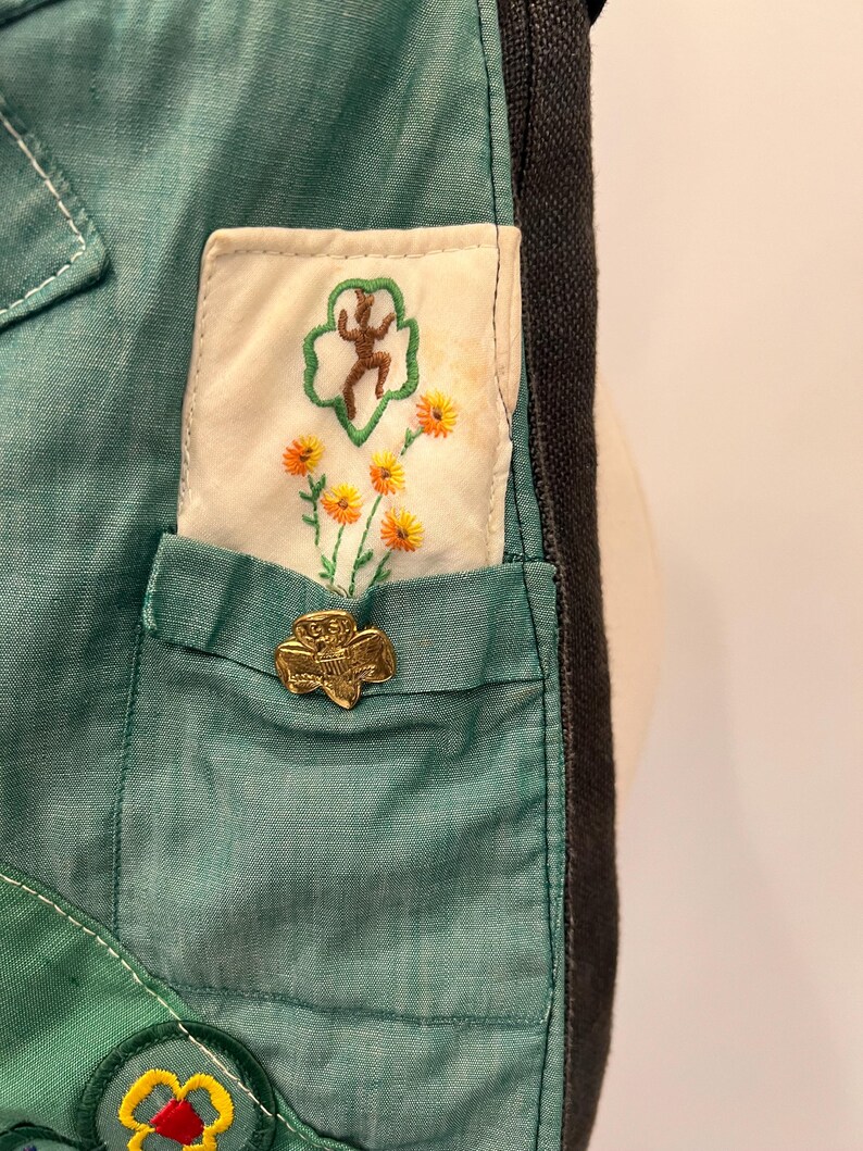 Puede incluir: Primer plano de una tela verde azulado con un bolsillo que presenta un alfiler dorado y un parche bordado con una figura y flores. El parche es blanco con un borde verde. Se ven costuras blancas.