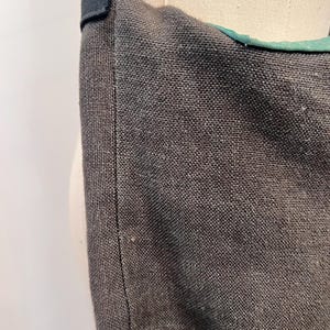 Puede incluir: Primer plano de un bolso tote de tela gris oscuro con una correa negra. El bolso tiene un tejido texturizado y un forro verde visible en el borde superior. La correa est&aacute; sujeta con una tela negra.