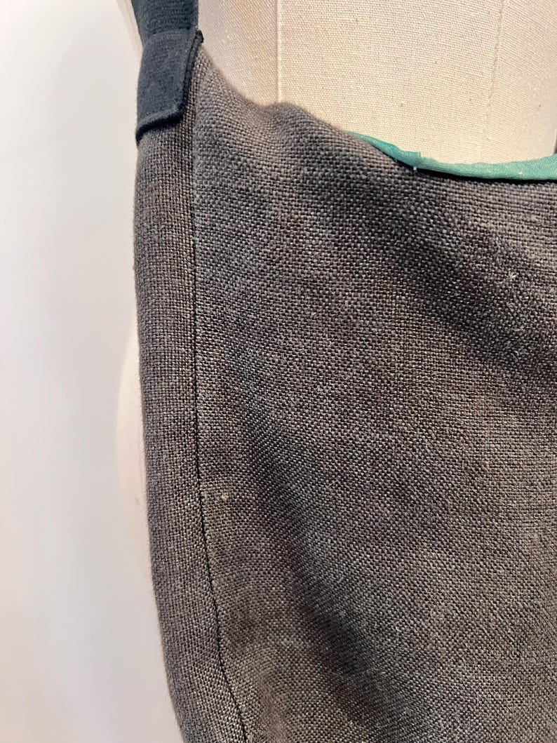 Puede incluir: Primer plano de un bolso tote de tela gris oscuro con una correa negra. El bolso tiene un tejido texturizado y un forro verde visible en el borde superior. La correa est&aacute; sujeta con una tela negra.