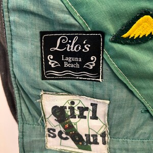 Puede incluir: Primer plano de un uniforme de Girl Scout verde y verde azulado. El uniforme presenta parches con el texto "Lilo's Laguna Beach" y "Girl Scout". Tambi&eacute;n son visibles un pin de estrella dorada y un parche de ala amarilla.