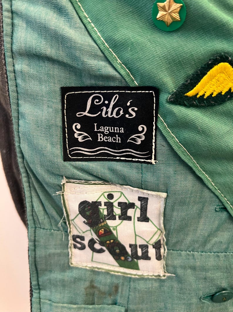 Puede incluir: Primer plano de un uniforme de Girl Scout verde y verde azulado. El uniforme presenta parches con el texto "Lilo's Laguna Beach" y "Girl Scout". Tambi&eacute;n son visibles un pin de estrella dorada y un parche de ala amarilla.