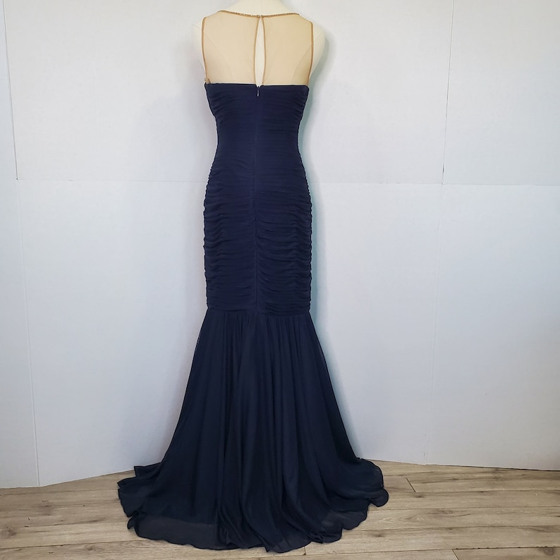 May include: A navy blue, floor-length, sleeveless gown with a fitted bodice and a flowing skirt. The gown has a sheer, nude-colored overlay at the neckline.