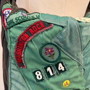 Puede incluir: Primer plano de un bolso verde y gris de Girl Scout con parches y detalles bordados. El bolso presenta las palabras "Girl Scouts" y "Washington Rock" en rojo y verde. Los parches incluyen una flor morada y los n&uacute;meros "814".