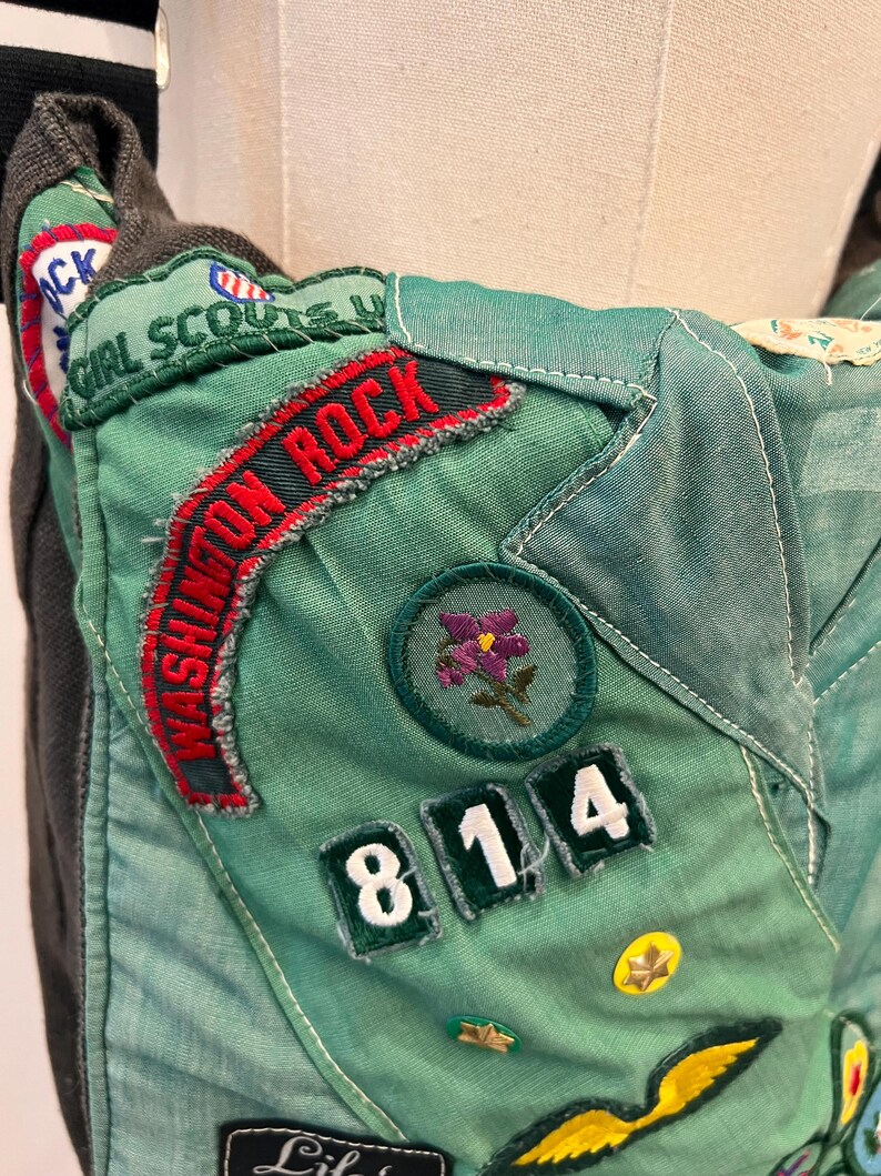 Puede incluir: Primer plano de un bolso verde y gris de Girl Scout con parches y detalles bordados. El bolso presenta las palabras "Girl Scouts" y "Washington Rock" en rojo y verde. Los parches incluyen una flor morada y los n&uacute;meros "814".