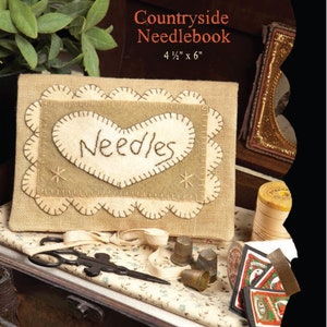 May include: A brown needlebook with a heart-shaped flap that says "Needles" in white stitching. The needlebook is 4 1/2 inches by 6 inches. The needlebook is sitting on a wooden surface with a pair of scissors, a thimble, and a spool of thread.