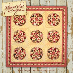 Pode incluir: Uma colcha com fundo amarelo e um design de patchwork vermelho, marrom e creme. A colcha é composta por nove quadrados, cada um com um design circular no centro. A colcha é cercada por uma borda vermelha e creme. O texto "Wagons West Designs" é visível no canto superior esquerdo da imagem.