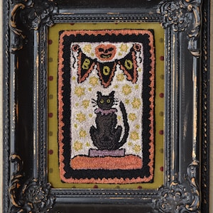 May include: A framed Halloween-themed textile art piece. The artwork features a black cat with a purple collar, sitting below a banner that reads "BOO" and a jack-o'-lantern. The frame is black with ornate detailing, and the background is a mix of green and orange.