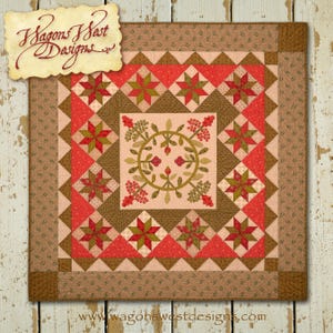 Pode incluir: Um padrão de colcha com um desenho central de uma coroa com bagas e folhas, rodeado por uma borda de estrelas vermelhas e marrons. A colcha é emoldurada por uma borda marrom com uma borda menor de padrão marrom e verde. O texto "Wagons West Designs" está no canto superior esquerdo e "www.wagonswestdesigns.com" está na parte inferior.