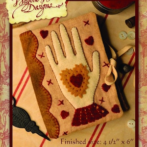 May include: A tan needlebook with a hand design, featuring a cream-colored hand with a heart-shaped center and red heart accents. The finished size is 4.5" x 6". The cover is embellished with a brown border and the text "Heart in Hand Needlebook".