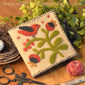 May include: A handmade felt needlepoint project featuring a red bird perched on a green branch with red berries and flowers. The project is framed with a brown stitched border and is displayed on a wooden surface.