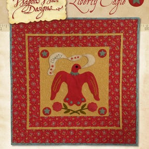 Pode incluir: Um bloco de colcha vermelho, branco e azul com uma águia vermelha estilizada com uma faixa que diz "Liberty" e uma estrela. O bloco é cercado por uma borda amarela e uma borda vermelha com um padrão de bolinhas brancas. O bloco de colcha mede 21 1/2 polegadas por 21 1/2 polegadas.