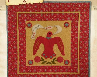 Puede incluir: Un bloque de colcha rojo, blanco y azul con un águila roja estilizada con una pancarta que dice "Liberty" y una estrella. El bloque está rodeado por un borde amarillo y un borde rojo con un patrón de lunares blancos. El bloque de colcha mide 21 1/2 pulgadas por 21 1/2 pulgadas.
