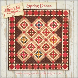 Pode incluir: Um padrão de colcha chamado "Spring Dance" de Wagons West Designs. A colcha é feita com uma variedade de tecidos em tons de vermelho, marrom, creme e azul. A colcha é emoldurada com uma borda marrom e vermelha.
