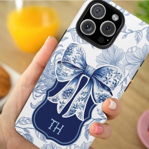 Puede incluir: Una funda de teléfono blanca con un diseño floral azul y blanco y un lazo azul. La funda tiene un monograma azul marino con las iniciales "TH".