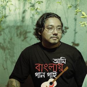 May include: A man wearing a black t-shirt with the text "আমি বাংলায় গান গাই" and a white acoustic guitar graphic.