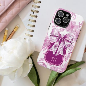 May include: A white phone case featuring a floral pattern and a large purple bow. The bow is set on a purple label with the initials "TH". The case is decorated with pink flowers and leaves.