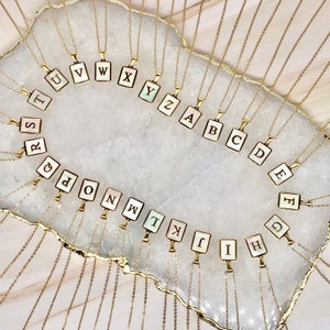 May include: A set of 26 gold necklaces with square pendants featuring each letter of the alphabet. The pendants are made of white mother-of-pearl with black lettering.