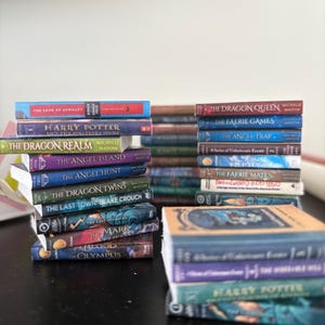 May include: Three stacks of various paperback and hardcover books with visible titles including The Song of Achilles, Harry Potter and the Sorcerer's Stone, The Dragon Realm, The Angel Island, The Angel Hunt, The Dragon Twins, and The Blood of Olympus.