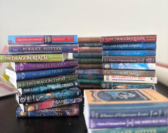 May include: Three stacks of various paperback and hardcover books with visible titles including The Song of Achilles, Harry Potter and the Sorcerer's Stone, The Dragon Realm, The Angel Island, The Angel Hunt, The Dragon Twins, and The Blood of Olympus.