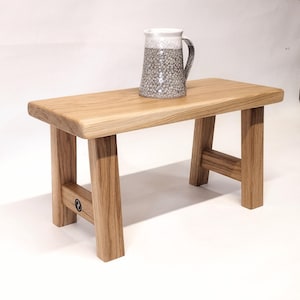 Op de afbeelding: Een kleine, rechthoekige houten bank met vier stevige poten. Een decoratieve keramische kan met een grijs en wit gespikkeld patroon staat op de bank. De bank is gemaakt van lichtgekleurd hout.