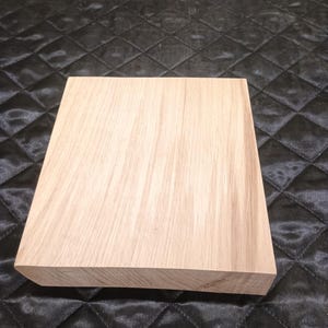 Può includere: Una tavola di legno rettangolare di colore chiaro, con venature visibili. La tavola ha una superficie liscia e non rifinita, con un bordo smussato. Il legno sembra essere rovere chiaro o un legno duro simile. Su uno sfondo scuro e trapuntato.