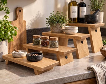 May include: A set of three light brown wooden risers with rectangular tops and angled legs, each featuring a small circular logo on the side. These tiered display stands are arranged to hold various kitchen items like spice jars and small bowls.