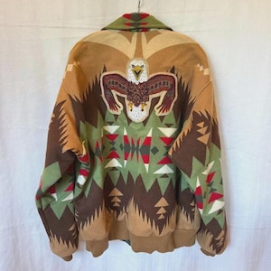 May include: A tan, brown, and green jacket featuring a large embroidered bald eagle on the back. The garment displays a geometric pattern in red, white, and black, with a fold-over collar and ribbed cuffs and hem, presented from the back view.