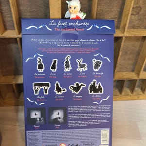 Peut inclure: Une bo&icirc;te bleue avec le titre "La for&ecirc;t enchant&eacute;e - The enchanted forest" et des illustrations de personnages de contes de f&eacute;es. Une petite figurine de f&eacute;e est pos&eacute;e dessus. La bo&icirc;te contient du texte en fran&ccedil;ais et en anglais.