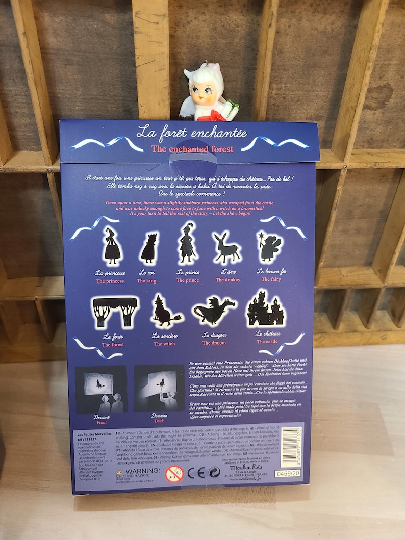 Peut inclure: Une bo&icirc;te bleue avec le titre "La for&ecirc;t enchant&eacute;e - The enchanted forest" et des illustrations de personnages de contes de f&eacute;es. Une petite figurine de f&eacute;e est pos&eacute;e dessus. La bo&icirc;te contient du texte en fran&ccedil;ais et en anglais.