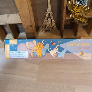 Peut inclure: Une bo&icirc;te de jouets pour enfants avec le texte "Les histoires du soir" et des illustrations d'animaux. La bo&icirc;te est bleue et porte une &eacute;tiquette d'avertissement. Une d&eacute;coration dor&eacute;e de la Tour Eiffel et un petit sapin de No&euml;l sont en arri&egrave;re-plan.