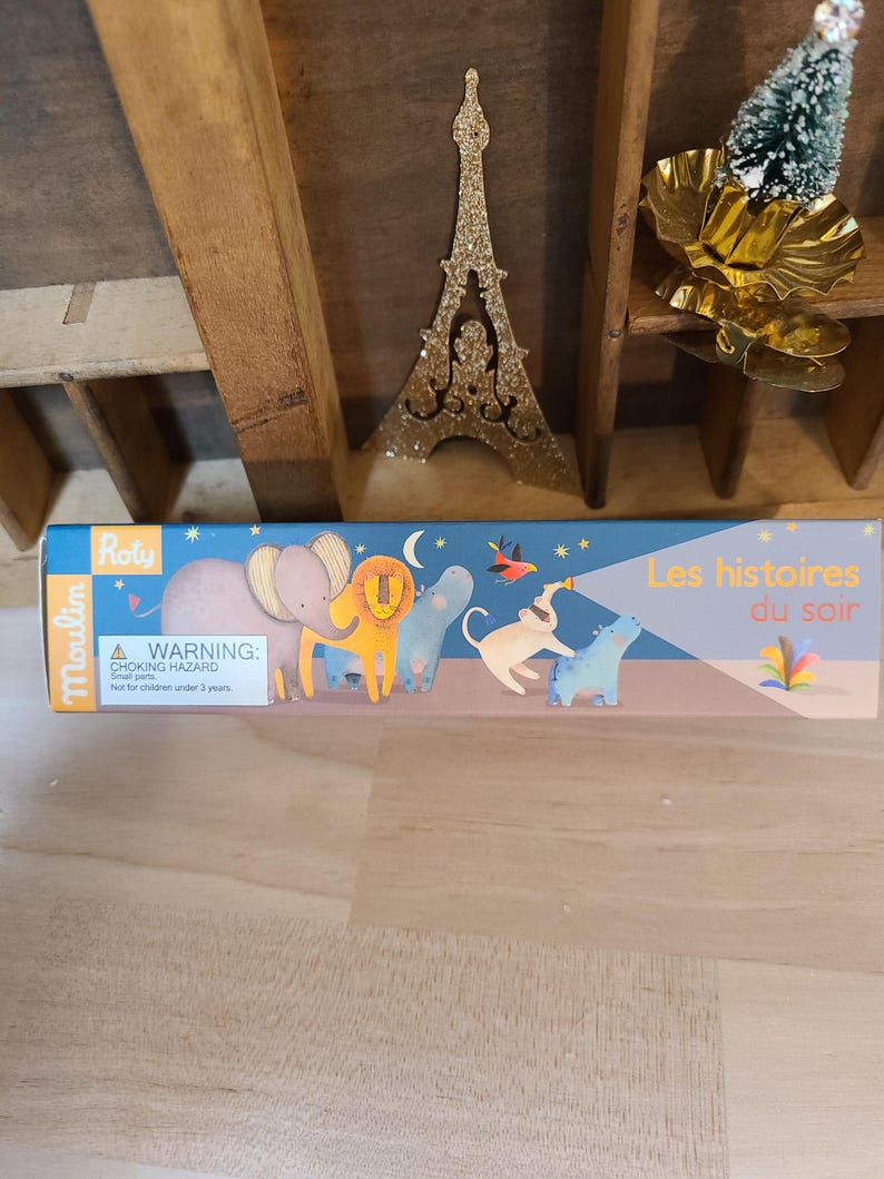 Peut inclure: Une bo&icirc;te de jouets pour enfants avec le texte "Les histoires du soir" et des illustrations d'animaux. La bo&icirc;te est bleue et porte une &eacute;tiquette d'avertissement. Une d&eacute;coration dor&eacute;e de la Tour Eiffel et un petit sapin de No&euml;l sont en arri&egrave;re-plan.