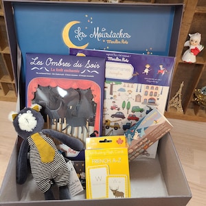 Peut inclure: Une bo&icirc;te grise remplie de jouets et de livres pour enfants. Comprend un chat en peluche, des cartes flash et des livres avec du texte en fran&ccedil;ais. Le couvercle de la bo&icirc;te pr&eacute;sente un croissant de lune et les mots "Les Moustaches" et "Moulin Roty".