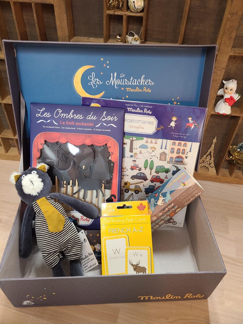 Peut inclure: Une bo&icirc;te grise remplie de jouets et de livres pour enfants. Comprend un chat en peluche, des cartes flash et des livres avec du texte en fran&ccedil;ais. Le couvercle de la bo&icirc;te pr&eacute;sente un croissant de lune et les mots "Les Moustaches" et "Moulin Roty".