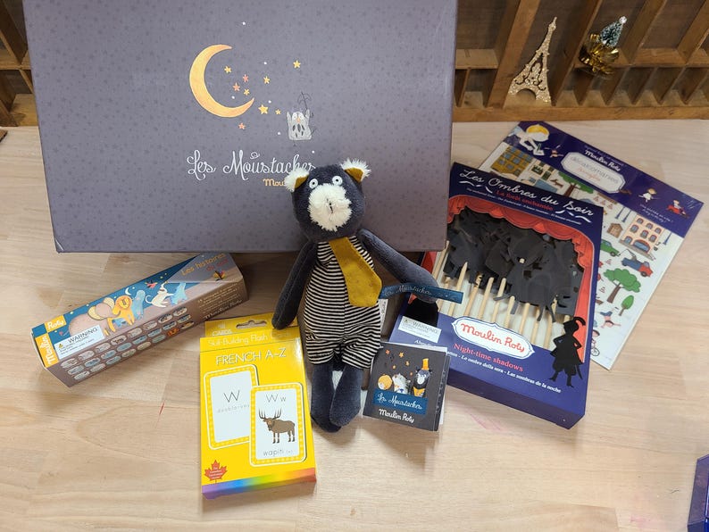 Peut inclure: Ensemble de jouets et de mat&eacute;riel &eacute;ducatif pour enfants. Comprend une peluche ours, des cartes de l'alphabet, des ombres chinoises et une bo&icirc;te avec un croissant de lune. Le texte "Les Moustaches" est visible sur la bo&icirc;te.
