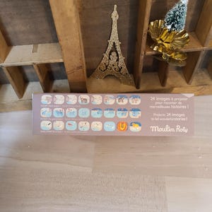 Peut inclure: Une bo&icirc;te rectangulaire avec le texte "24 images &agrave; projeter pour raconter de merveilleuses histoires!" et "Moulin Roty". La bo&icirc;te pr&eacute;sente des illustrations d'animaux et de sc&egrave;nes. Une d&eacute;coration dor&eacute;e de la Tour Eiffel et un petit sapin de No&euml;l sont en arri&egrave;re-plan.