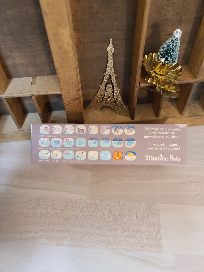 Peut inclure: Une bo&icirc;te rectangulaire avec le texte "24 images &agrave; projeter pour raconter de merveilleuses histoires!" et "Moulin Roty". La bo&icirc;te pr&eacute;sente des illustrations d'animaux et de sc&egrave;nes. Une d&eacute;coration dor&eacute;e de la Tour Eiffel et un petit sapin de No&euml;l sont en arri&egrave;re-plan.