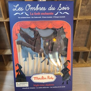 Peut inclure: Un coffret de jeu "Les Ombres du Soir" de Moulin Roty. La bo&icirc;te est bleu fonc&eacute; avec des accents rouges et blancs, avec le texte "La for&ecirc;t enchant&eacute;e" et "Night-time shadows". &Agrave; l'int&eacute;rieur, des silhouettes cr&eacute;ent une sc&egrave;ne de for&ecirc;t.
