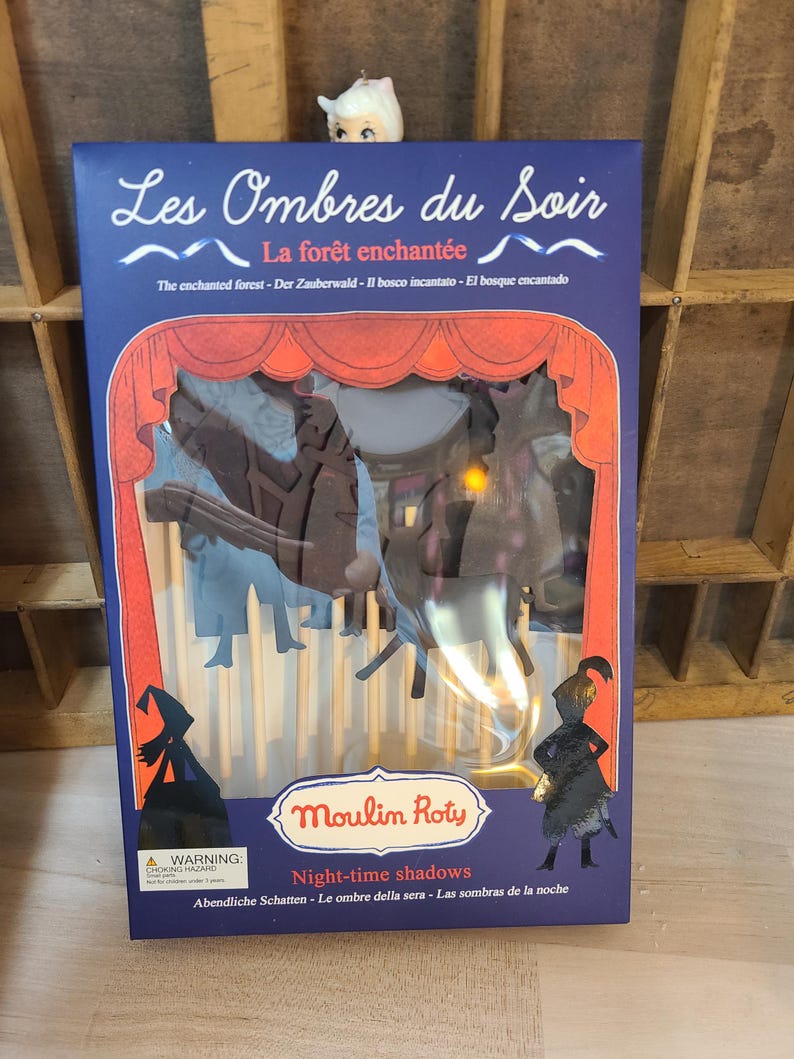 Peut inclure: Un coffret de jeu "Les Ombres du Soir" de Moulin Roty. La bo&icirc;te est bleu fonc&eacute; avec des accents rouges et blancs, avec le texte "La for&ecirc;t enchant&eacute;e" et "Night-time shadows". &Agrave; l'int&eacute;rieur, des silhouettes cr&eacute;ent une sc&egrave;ne de for&ecirc;t.