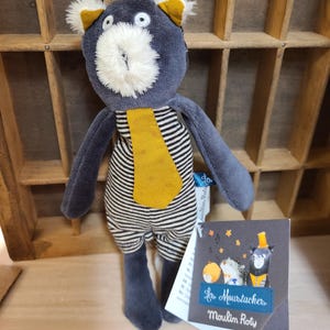 Peut inclure: Poup&eacute;e ours en peluche bleu marine avec un corps ray&eacute; noir et blanc, une cravate et des oreilles jaunes, et un museau blanc duveteux. L'&eacute;tiquette indique "Les Moustaches Moulin Roty".