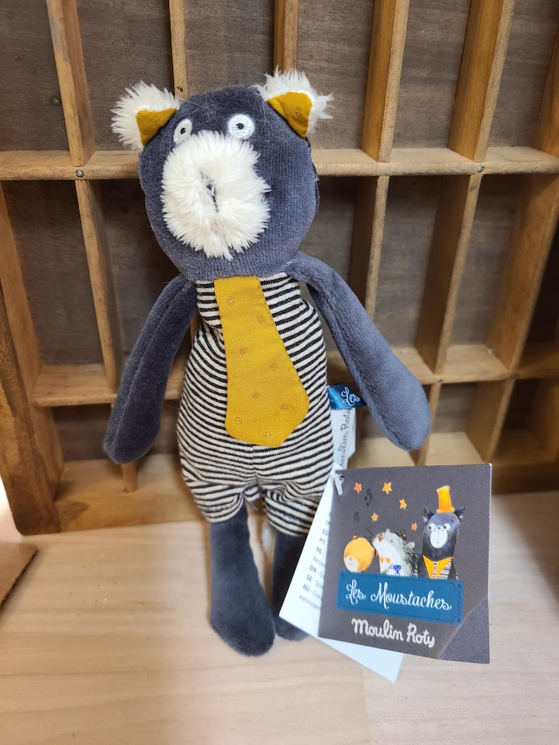Peut inclure: Poup&eacute;e ours en peluche bleu marine avec un corps ray&eacute; noir et blanc, une cravate et des oreilles jaunes, et un museau blanc duveteux. L'&eacute;tiquette indique "Les Moustaches Moulin Roty".