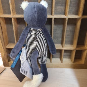 Peut inclure: Un chat en peluche avec un corps gris, des oreilles blanches et une barboteuse &agrave; rayures noires et blanches. Le chat a une queue blanche duveteuse et tient une &eacute;tiquette.