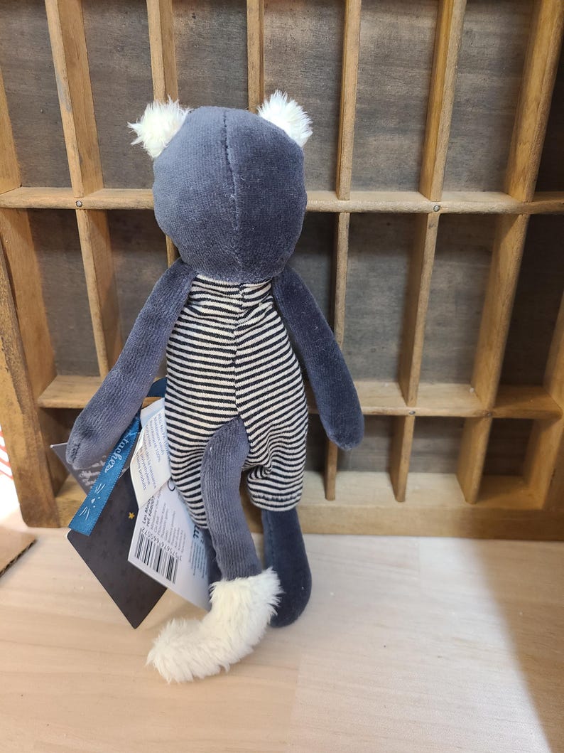 Peut inclure: Un chat en peluche avec un corps gris, des oreilles blanches et une barboteuse &agrave; rayures noires et blanches. Le chat a une queue blanche duveteuse et tient une &eacute;tiquette.