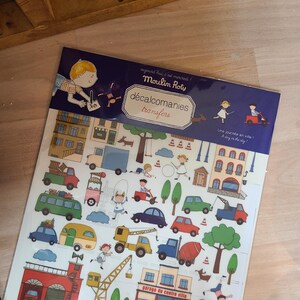 Peut inclure: Une planche de d&eacute;calcomanies "Moulin Roty" sur le th&egrave;me de la ville. La planche comprend des illustrations de voitures, de b&acirc;timents, d'arbres et d'enfants. Le texte sur la planche indique "d&eacute;calcomanies transfers" et "Une journ&eacute;e en ville!"