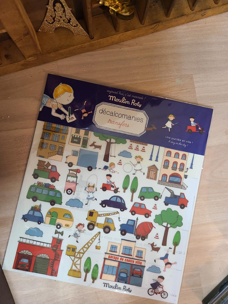 Peut inclure: Une planche de d&eacute;calcomanies "Moulin Roty" sur le th&egrave;me de la ville. La planche comprend des illustrations de voitures, de b&acirc;timents, d'arbres et d'enfants. Le texte sur la planche indique "d&eacute;calcomanies transfers" et "Une journ&eacute;e en ville!"