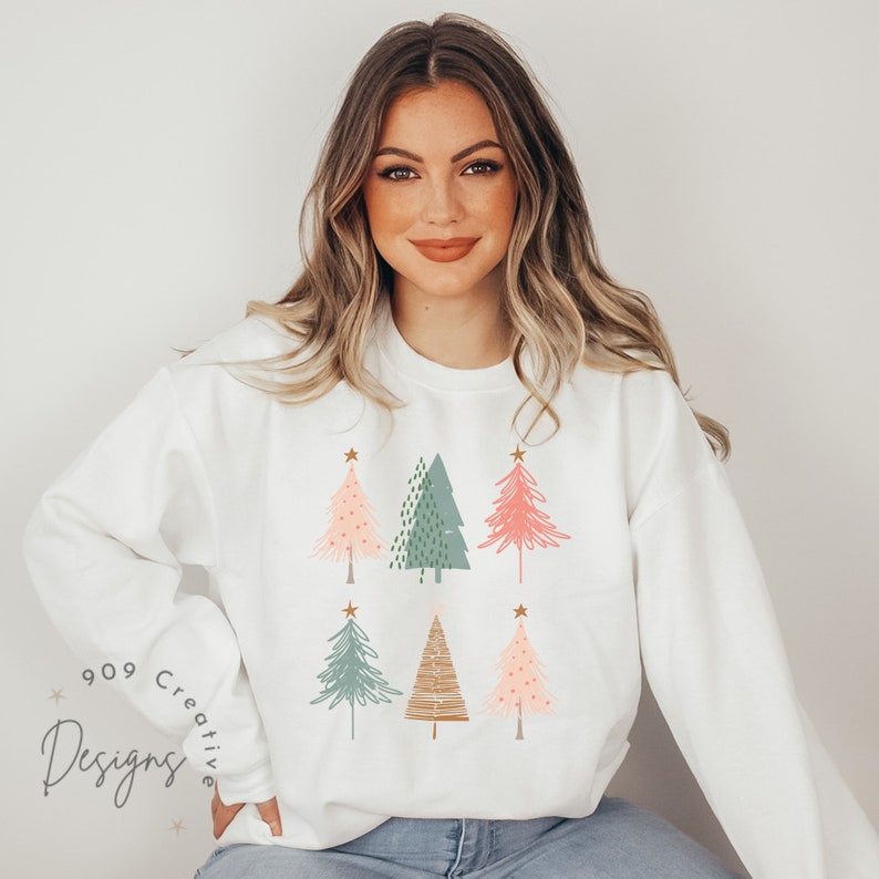 May include: White sweatshirt featuring a festive design of six stylised Christmas trees in shades of green, pink, and brown. Each tree has a gold star on top.