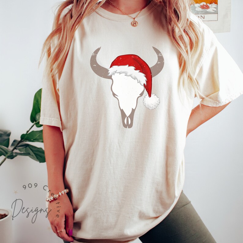 May include: A white t-shirt featuring a graphic of a bull skull wearing a red Santa hat. The shirt is a light cream colour and has a relaxed fit.