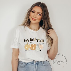 Puede incluir: Camiseta blanca con el texto "it's fall y'all" en negro con una fuente retro. La camiseta presenta un gr&aacute;fico de cinco calabazas en varios colores y patrones con estrellas negras.