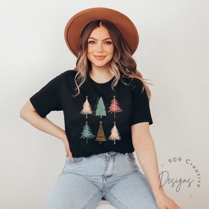 May include: A black t-shirt featuring a graphic of six colourful Christmas trees. The trees are stylised and have a retro look. The shirt is being worn by a person with brown hair and a brown hat.